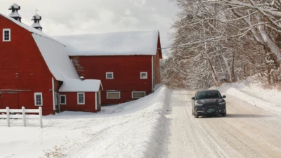An electric vehicle driving on a dirt road covered with snow, passing by a red barn.
